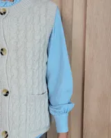 Chaleco de punto con diseño de trenzas, color gris oscuro, con cierre frontal de botones grandes y bolsillos laterales.