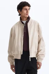 Campera bomber en mezcla de algodón y nylon color beige, con cuello elevado acanalado y cierre frontal de cremallera. Cuenta con bolsillos laterales y un bolsillo con cierre en la manga izquierda. Puños y bajo en punto acanalado, de corte holgado.