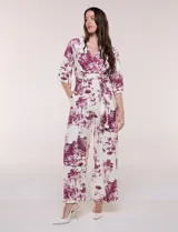 Enterito largo de calce fluido con escote en V cruzado, mangas 3/4 anchas, cintura elastizada con lazo ajustable y pantalón wide leg con bolsillos laterales. Estampado tipo toile de Jouy en tonos violeta sobre fondo blanco.