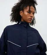 Campera rompevientos azul marino con cuello alto ajustable, cierre frontal y cordones en la cintura para un ajuste entallado. Presenta detalles de costuras en contraste blanco y puños elásticos.