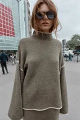 Jersey de punto con cuello subido y manga larga, de corte holgado. Presenta detalles de vivos en color crema en el cuello, puños y costuras de las mangas, contrastando con el tono marrón jaspeado de la prenda.