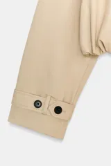 Trench corto de color beige con cuello de solapa y escote en pico. Presenta un diseño de cierre frontal cruzado con botones, mangas largas con trabillas en los puños y un bajo con acabado abullonado gracias a un elástico interior. Incluye bolsillos laterales ocultos y forro interior.