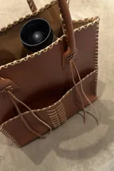 Bolso tipo tote de cuero vacuno color suela, confeccionado artesanalmente con costuras decorativas visibles en los bordes y en el centro. Cuenta con doble asa larga para llevar al hombro y detalles de tiras colgantes en la unión de las asas.