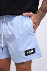 Short de baño color celeste, con cordón ajustable en la cintura y un parche cuadrado negro con el logo "HAUS" en blanco en la pierna izquierda.