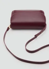 Bolso tipo bandolera de tamaño pequeño con acabado efecto piel en color bordó. Presenta una solapa frontal con cierre metálico a presión, costuras visibles en contraste y una correa larga ajustable con herrajes dorados.