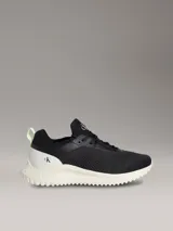 Zapatillas tipo runner Calvin Klein en color negro con suela de EVA blanca y detalles en verde claro en el talón. Presentan diseño tipo calcetín, parte superior de malla técnica y logo de la marca en la lengüeta y el lateral.