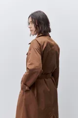 Trench clásico color marrón, de silueta relajada, confeccionado en tejido liviano, con cuello de solapa, doble abotonadura frontal y mangas largas con presillas regulables en los puños.