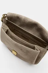 Bolso bandolera de piel gamuzada color gris topo, con solapa curva, asa de mano corta y herrajes metálicos dorados.