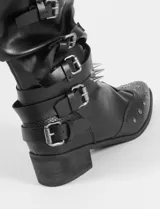 Botas de caña media en color negro, confeccionadas en cuero sintético. Presentan un diseño con múltiples correas con hebillas metálicas, tachas decorativas en la puntera y una tira con tachas cónicas alrededor del tobillo. Incluyen un taco bajo cuadrado y suela de caucho.