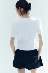 Blusa corta blanca con escote en U, mangas cortas abullonadas y cierre frontal con botones negros contrastantes.