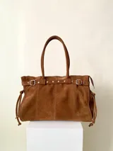 Bolso grande de cuero marrón con textura tipo cocodrilo, estructura rígida y asas para llevar al hombro. Tiene una tira con tachas y hebillas en cada extremo, también lleva lazos en los costados.