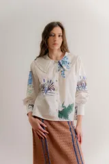 Camisa de popelina de algodón color crudo, con cuello bobo amplio con volados y cierre frontal de botones. Presenta un diseño estampado con motivos botánicos y de aves en tonos azules y verdes.