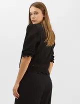 Blusa de algodón semitransparente con diseño floral bordado a tono. Presenta escote en V, cierre frontal con botones y mangas al codo ligeramente abullonadas. Incluye detalles de encaje calado estilo macramé en los puños y en el ruedo con terminaciones en ondas.