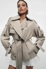 Trench corto de corte oversize confeccionado en mezcla de algodón con acabado repelente al agua. Presenta cuello solapa, cierre frontal con botones, mangas largas con trabillas ajustables y cinturón a tono con lazada en la cintura.