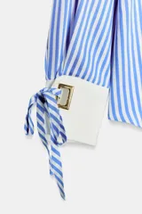 Camisa de cuello solapa y manga larga con rayas verticales celestes y blancas. Cuenta con puños anchos blancos contrastantes, ajustables con hebilla dorada y lazos, y cierre frontal con botones dorados.