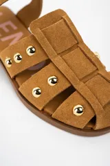 Sandalia tipo cangrejera de gamuza color camel, con diseño de tiras entrelazadas y tachas metálicas doradas. Cuenta con cierre de pulsera con hebilla ajustable y suela plana de goma.