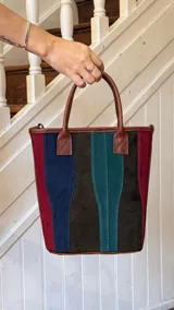 Matera de cuero con diseño de paneles verticales en combinación de colores azul, verde, marrón y bordó. Cuenta con dos asas cortas de cuero y estructura rígida.