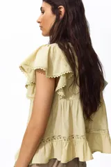 Blusa de tejido liviano color amarillo claro, con escote en V profundo, mangas cortas estilo mariposa y cierre frontal con botones. Presenta detalles de alforzas e inserciones de encaje en el frente y en el borde del ruedo y las mangas, terminando en un volante inferior.
