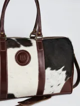 Bolso de viaje tipo duffel de cuero con detalles en pelo vacuno con estampado animal print blanco y negro. Cuenta con doble asa de mano, correa ajustable y desmontable, y detalles en marrón oscuro.