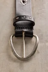Cinturón de cuero marrón con calados en forma de gota y tachas metálicas a lo largo de la tira. Cuenta con hebilla metálica ovalada y ajuste regulable.