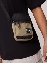 Bandolera negra de poliéster con logo de la marca Rusty en relieve en el frente. Cuenta con un bolsillo frontal con solapa y cierre de cremallera en el compartimento principal. La correa es ajustable.