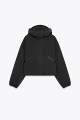 Anorak corto de color negro con acolchado interior, cuello subido y capucha. Presenta mangas largas con puños elásticos, bolsillos delanteros con cierre y bajo ajustable con cordones. Cierre frontal con cremallera termosellada y botones a presión.