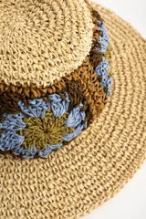 Sombrero de ala ancha color tostado, tejido en papel con detalles de apliques de crochet en la parte central con diseño de flores en tonos celestes, marrones y verde oliva.