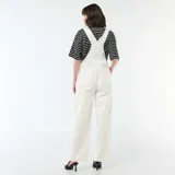 Enterito de jean color blanco con diseño de pierna ancha, bolsillo frontal en el pecho y tirantes ajustables con hebillas metálicas.
