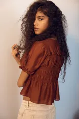 Blusa marrón de lyocell con escote redondo, mangas cortas abullonadas y cuerpo fruncido con detalle de tiras decorativas.