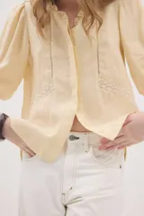 Camisa de lino color beige, de silueta romántica con escote a la base y mini volado. Cuenta con mangas 3/4 abullonadas con frunces y terminación en elástico. Posee detalles de puntillas negras en contraste en el frente y cierre frontal con botones ocultos bajo cartera.
