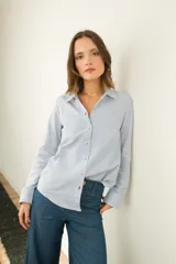 Camisa celeste a rayas verticales blancas, de corte clásico con cuello camisero, mangas largas y cierre frontal con botones rojos. Presenta costuras en contraste y un corazón bordado en la espalda.