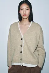 Chaqueta tipo cárdigan de lana en color beige, con diseño de tejido combinado que incluye una capa interior de tela clara. Presenta escote en pico, manga larga y cierre frontal con botones oscuros.