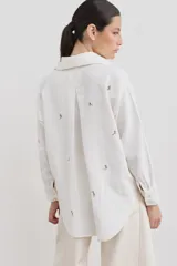 Camisa de corte holgado color blanco, con cuello clásico, cierre frontal de botones y bolsillo delantero. Presenta un delicado bordado de figuras deportivas en color marrón.