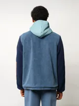 Campera de polar con capucha, cierre frontal completo y diseño en bloques de color azul y azul marino. Presenta detalles en nylon en la zona del cierre y la capucha, con logo de la marca aplicado en el pecho.