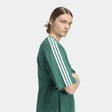 Remera Adidas de corte holgado en color verde, con cuello redondo, mangas cortas con las icónicas tres rayas blancas en los hombros y logo del trébol bordado en el pecho.