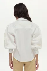 Camisa blanca de algodón con mangas abullonadas, cuello camisero y bordados geométricos y florales en tonos celestes y amarillos en el frente y las mangas.