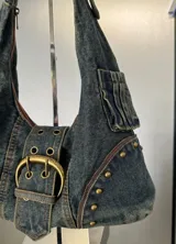 Cartera bandolera de denim azul con detalles de tachas y hebilla metálica.