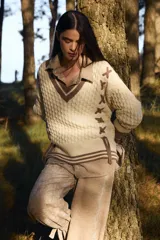 Sweater de punto estilo preppy en color crudo con detalles en marrón. Presenta cuello en V con ribete a rayas, tejido con textura de ochos y cordones laterales decorativos. Posee puños y ruedo acanalados con diseño a rayas.