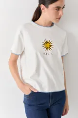 Remera blanca de algodón con cuello redondo y mangas cortas. Presenta un bordado central de un sol amarillo con la palabra 'SAVIA' debajo, y detalles de costura decorativa en color celeste en el cuello, mangas y ruedo.