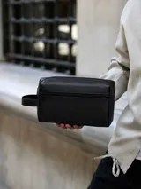 Bolso de mano tipo neceser en color negro, confeccionado en ecocuero con diseño minimalista, cierre principal superior y bolsillo frontal con cierre, incluye una manija lateral para transporte.