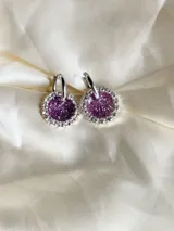 Aros colgantes con forma circular, hechos de resina color lila y borde de strass plateado. El aro es de acero quirúrgico plateado.