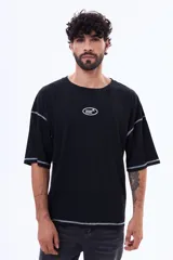 Remera negra de corte boxy con costuras a la vista en color blanco. Presenta un pequeño logo bordado en el centro del pecho y mangas cortas amplias.