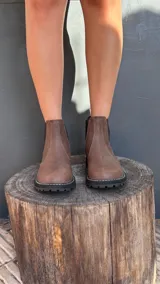 Bota tipo Chelsea de cuero marrón con textura granulada, paneles laterales elásticos en color negro y suela track dentada con costura blanca visible.