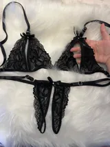 Conjunto de lencería de dos piezas confeccionado en encaje negro, compuesto por un soutiene triangular con aberturas frontales y una bombacha tipo colaless abierta con detalles de moños y apliques de strass.