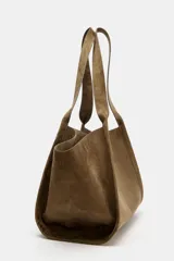 Cartera shopper de hombro confeccionada en gamuja color marrón, con diseño estructurado de laterales abiertos y doble asa superior. Cuenta con cierre de botón imantado e incluye un bolsillo interior extraíble con cremallera.