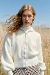 Chaqueta de punto color blanco con cuello subido, cierre frontal de cremallera metálica y mangas largas abullonadas con puños ajustados.