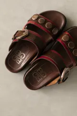 Sandalias bajas de cuero marrón con doble tira, hebilla ajustable y apliques de tachas metálicas.