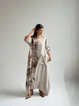 Vestido largo beige de lino con diseño midi holgado, cuello redondo, mangas cortas con vuelta y un gran estampado lateral de flores y hojas en tonos marrones y verdes. Cuenta con un bolsillo frontal aplicado con detalles deshilachados.