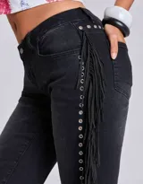 Pantalón de jean negro de corte acampanado, con detalle de flecos largos a lo largo de las costuras laterales y ojales metálicos decorativos.