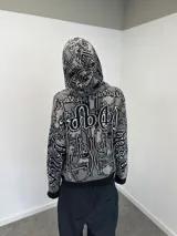 Buzo con cierre frontal completo, confeccionado en tejido de punto grueso de algodón. Presenta un diseño estampado integral con motivos gráficos y tipografía estilo gótico en tonos gris y negro, capucha integrada, puños y ruedo acanalados en color negro sólido.